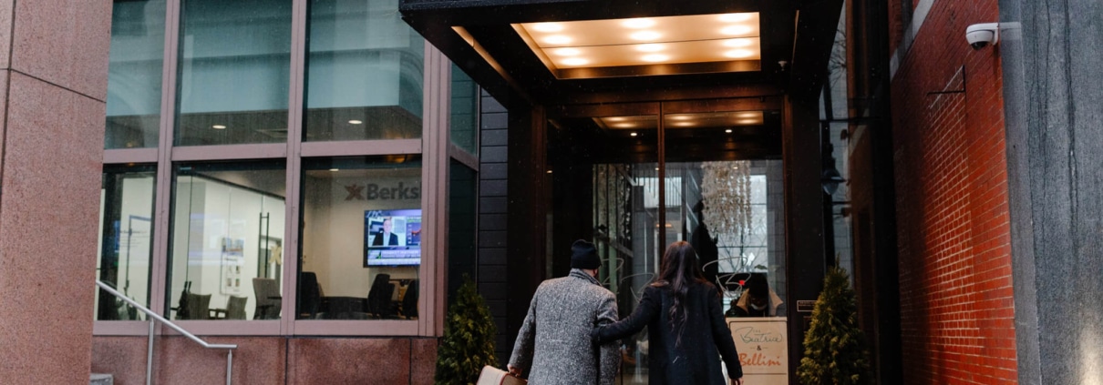 Two people in coats and hats walk toward the entrance of The Beatrice hotel on a wet city sidewalk, one carrying luggage, showcasing the impeccable hospitality management that defines the hotel's esteemed reputation in the industry.