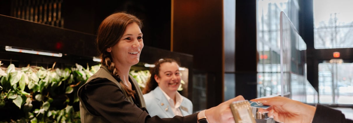 Two women stand at a counter, one woman smiles while handing an item to a person off-camera. The scene, reminiscent of the hospitality industry, features green plants and large windows bathed in natural light.