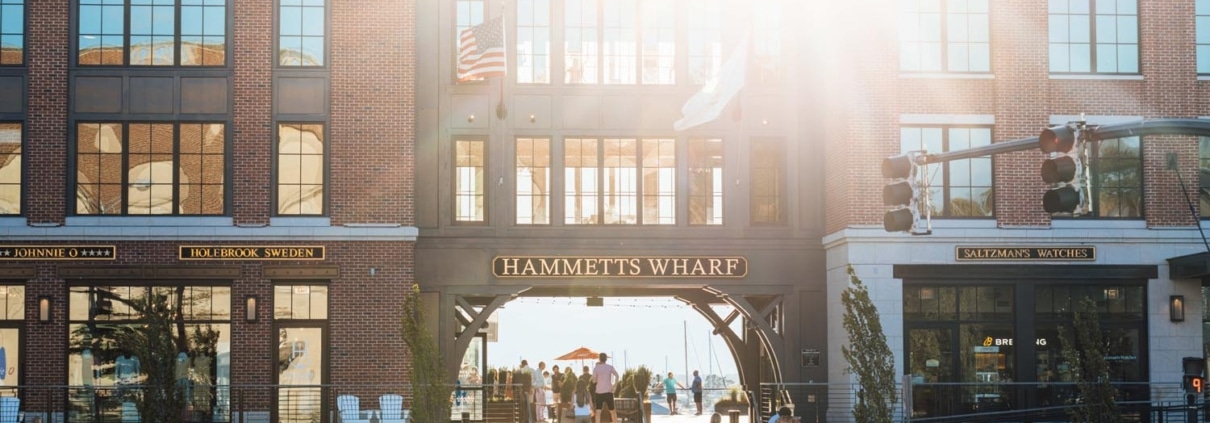 A sunlit scene of Hammett's Wharf, featuring a brick building with retail shops and people walking along the stairs and sidewalk. Reflecting the charm of the hospitality industry, American flags are displayed, and cars are visible on the street.