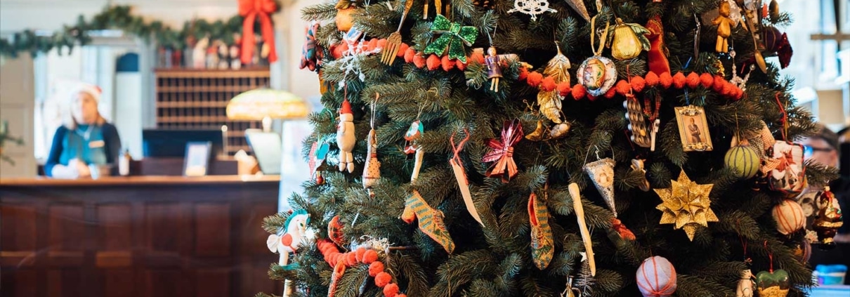 A large Christmas tree adorned with various ornaments and red garlands stands in a festively decorated hotel lobby, showcasing the warmth and charm synonymous with the hospitality industry, with a counter and a person in the background.