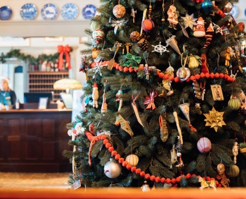 A large Christmas tree adorned with various ornaments and red garlands stands in a festively decorated hotel lobby, showcasing the warmth and charm synonymous with the hospitality industry, with a counter and a person in the background.