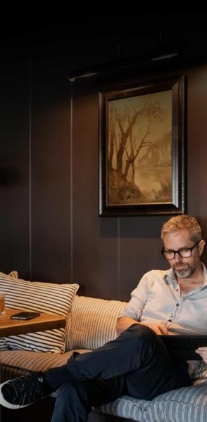 A man with glasses and a beard sits on a striped couch, reading a book. Behind him, on the dark wall, hangs a framed painting of trees—a tranquil scene reminiscent of the hospitality industry's calming ambiance. A nearby table holds a phone and a glass of water.