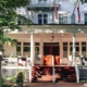 A large white house with a red door and front porch, adorned with rocking chairs and potted plants, flanked by trees. Two flags are mounted on the porch roof, reflecting its charm often celebrated in the hospitality industry. Red steps lead up to the welcoming entrance.