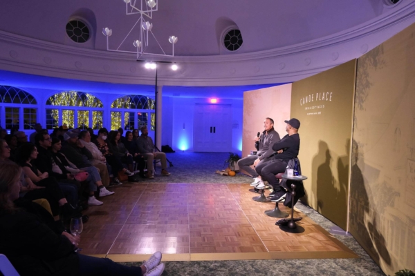 Three people sit on stools speaking into microphones on a stage in front of an audience in a dimly lit room with arched windows and a high ceiling. The backdrop reads "GRAVEL PLACE" and "KAMWE MAKE OVERS" as they discuss trends in the hospitality management industry.